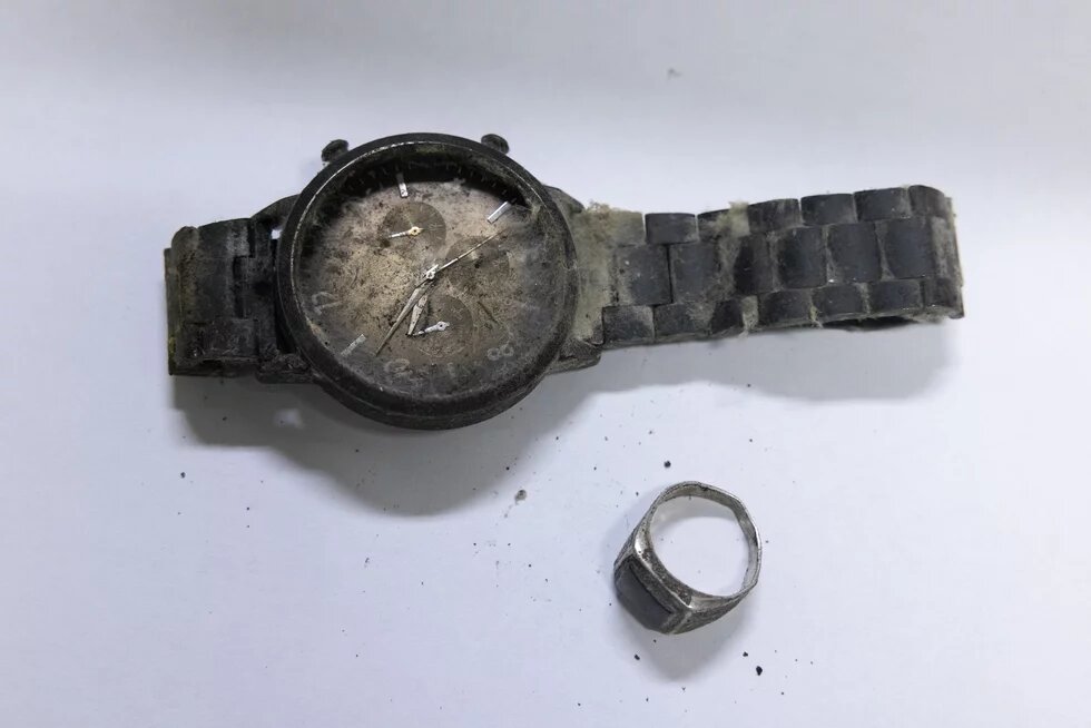 Eine stark verschmutzte, beschädigte Armbanduhr und ein verstaubter Ring liegen auf einer weißen Oberfläche.