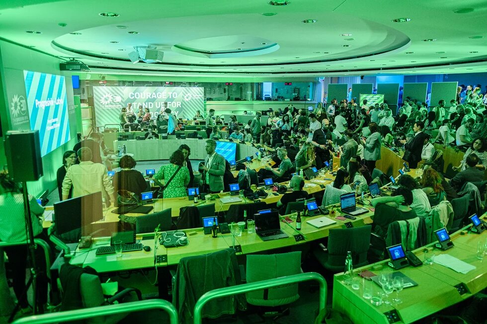 Ein Konferenzraum voller Teilnehmer*innen. Auf der Leinwand steht „Courage to Stand Up for Democracy“. Laptops und Mikrofone sind auf den Tischen.