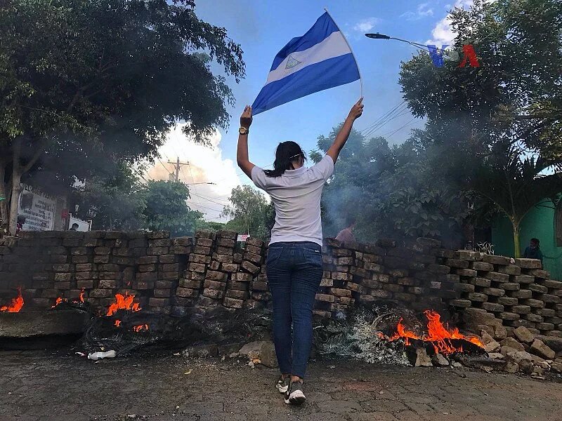 Eine Frau steht nahe einer brennenden Barrikade und hält die Staatsflagge von Nicaragua.