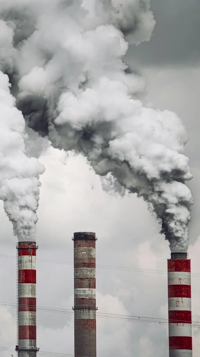 Foto: Mehrere Industrieschornsteine stoßen dicke, graue Rauchwolken aus. Stromleitungen im Hintergrund.