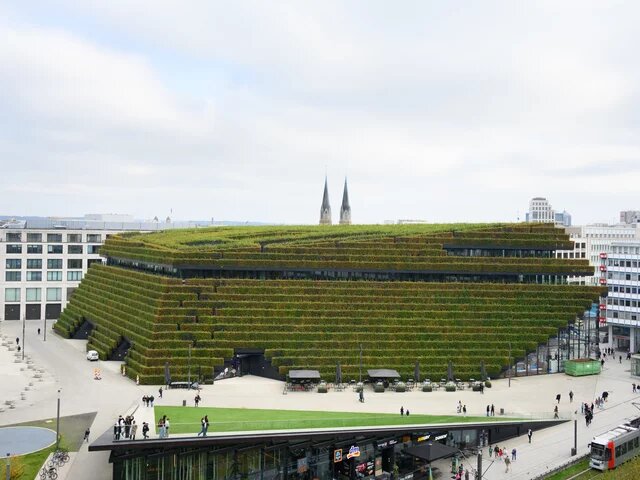  Gebäude mit terrassenförmig bepflanzter Fassade. Umgeben von städtischem Platz, dahinter moderne Gebäude und Kirchtürme.