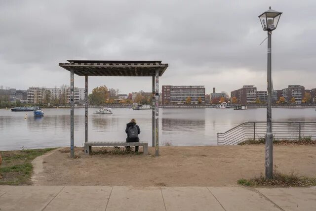 Person sitzt unter einem überdachten Sitzplatz am Wasser, blickt auf Boote und moderne Wohngebäude am gegenüberliegenden Ufer.