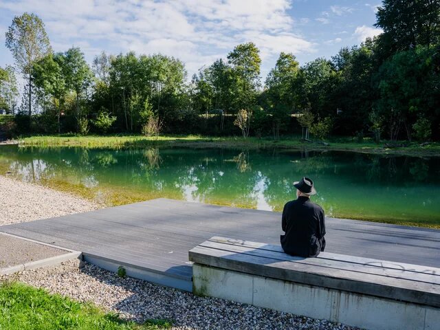 Eine Person sitzt auf einer Holzplattform an einem kleinen See mit grünem Wasser, umgeben von Bäumen und Kieselstrand.