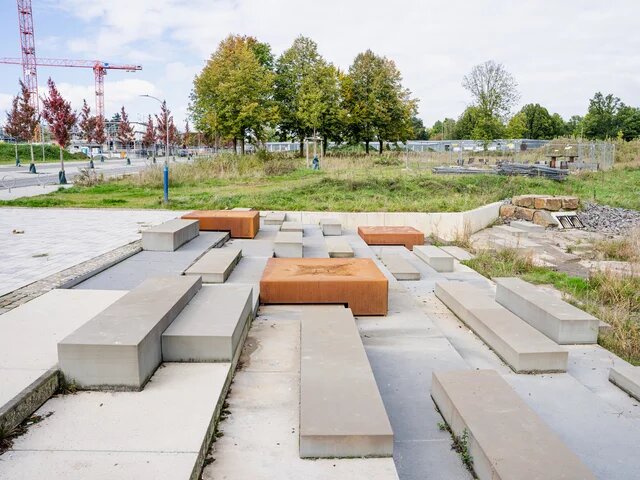 Rechteckige Beton- und Rostblöcke in einem offenen Bereich, vermutlich als Sitzflächen. Im Hintergrund Baustelle mit Kränen und Bäumen.