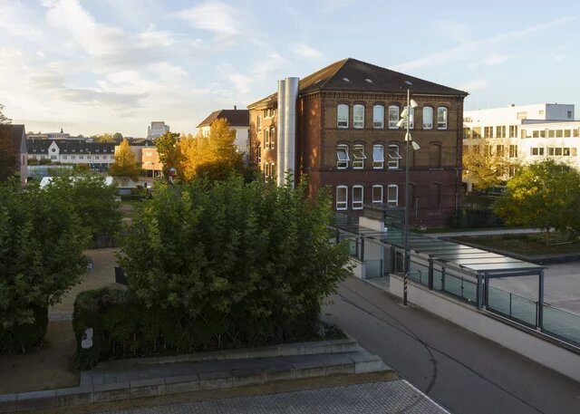 Historisches Backsteingebäude neben modernen Bauten, umgeben von Bäumen im Herbstlaub. Helle, teils bewölkte Abendstimmung.