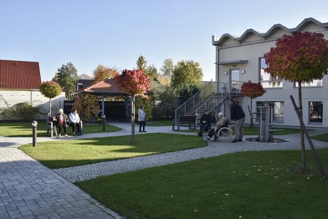 Gartenanlage mit Bäumen und Wegen, Menschen auf Bänken und im Rollstuhl. Im Hintergrund ein Pavillon und ein Gebäude mit Außentreppe.