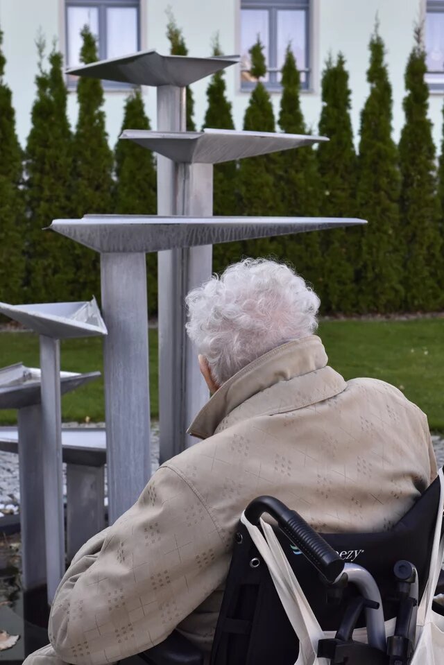Eine Person im Rollstuhl betrachtet eine moderne Metallskulptur im Freien. Im Hintergrund sind Hecken und Fenster eines Gebäudes zu sehen.