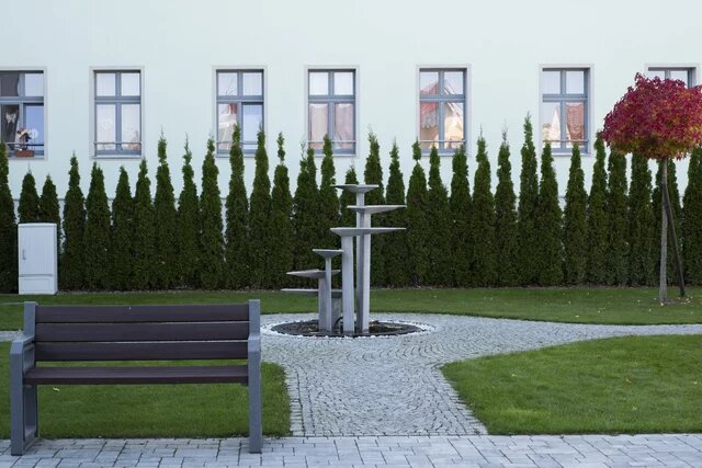 Garten mit Bank, Kiesweg und moderner Metallskulptur. Im Hintergrund Hecken und eine helle Gebäudeaußenwand mit Fenstern.