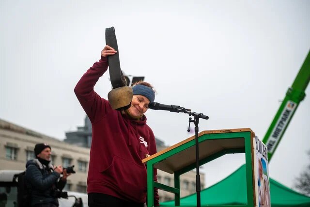Person am Rednerpult hebt Kuhglocke neben Mikrofon; im Hintergrund Gebäude, Zelt und Person mit Kamera.
