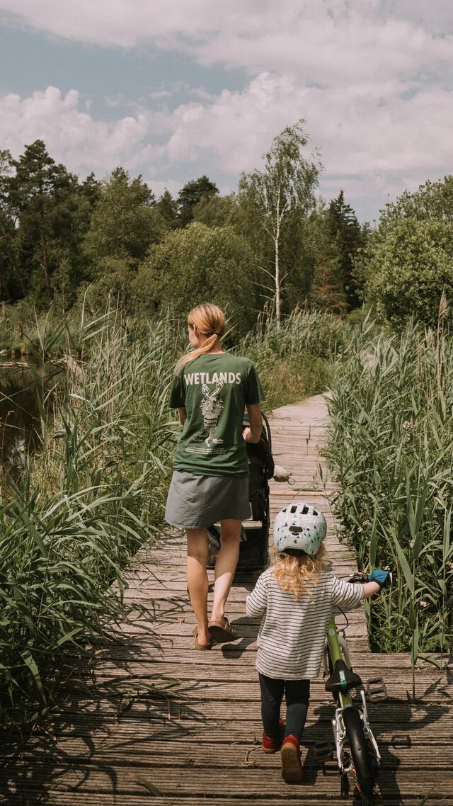 Eine Frau und ein Kind mit Laufrad gehen über einen Holzsteg durch dichtes Schilf an einem Teich.