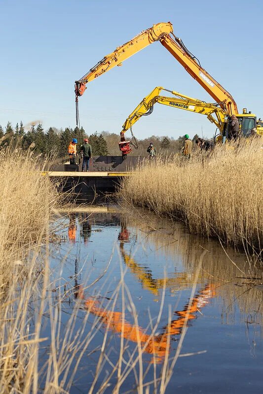 Zwei Bagger arbeiten an einer kleinen Brücke über einem schilfgesäumten Gewässer, während mehrere Personen die Arbeiten begleiten.