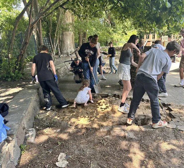 Eine Gruppe Kinder und Jugendlicher gräbt mit Schaufeln und Werkzeugen einen gepflasterten Bereich unter Bäumen auf.