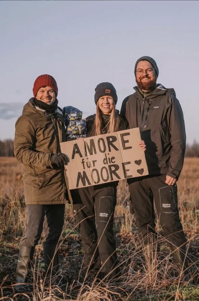 Drei Personen stehen lächelnd im Moor und halten ein Schild mit der Aufschrift „AMORE für die MOORE“ und zwei Herzen.