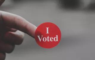 One hand is holding a sticker with "I Voted" written on it against a blurred background.