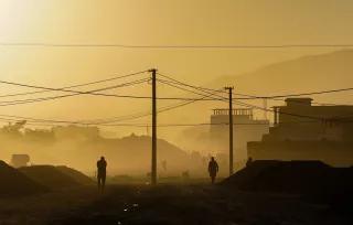 Schwarze Umrisse von zwei Menschen und Strommasten auf einer Straße, dahinter Kabul im gelben Nebel bei Sonnenaufgang