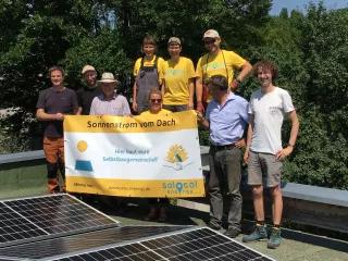 Menschen stehen vor Solarpanelen und halten ein Transparent von SoLocal Energy e.V. in der Hand