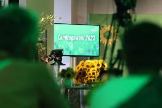 Ein großer Bildschirm zeigt eine grüne Wiese und darauf den Schriftzug "Landtagwahl 2023", davor liegen ein großer Strauß Sonnenblummen, im Hintergrund stehen Aufsteller der Partei Bündnis 90/Die Grünen. Im Vordergrund sind schemenhaft Personen mit Kameras zu erkennen.