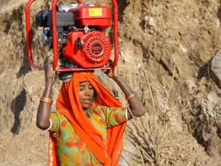 The women of the mountain villages in Rajasthan, India, carry 70 lbs. (or 32kgs) of wood on their heads for cooking every day. This woman is carrying a generator for repair in a nearby village