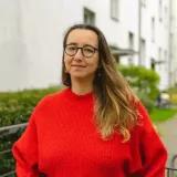 Porträt von Oleksandra Bienert. Sie trägt einen roten Pullover, lange, dunkelblonde Haare und eine Brille