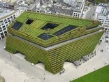 Begrüntes Gebäude mit Terrassenpflanzung. Dach mit offenen Innenhöfen, umgeben von städtischer Bebauung und Gehwegen.