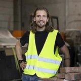 Person mit langen Haaren und Warnweste steht in Werkhalle neben einem Stapel Ziegelsteine.