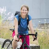 Eine Person auf einem Fahrrad, im Hintergrund eine Wiese und ein Gebäude.
