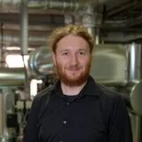 Person mit Bart steht vor metallischen Rohrleitungen in einem Technikraum und blickt in die Kamera.