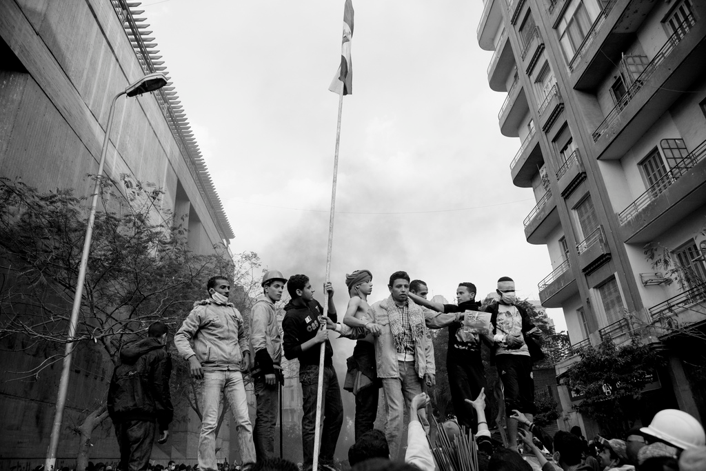 Eine Gruppe junger Männer in Ägypten Eine Gruppe junger Männer steht erhöht zwischen zwei Häuserblocks über einer Menschenmenge. Einer hält eine eingerollte Fahne in die Höhe.